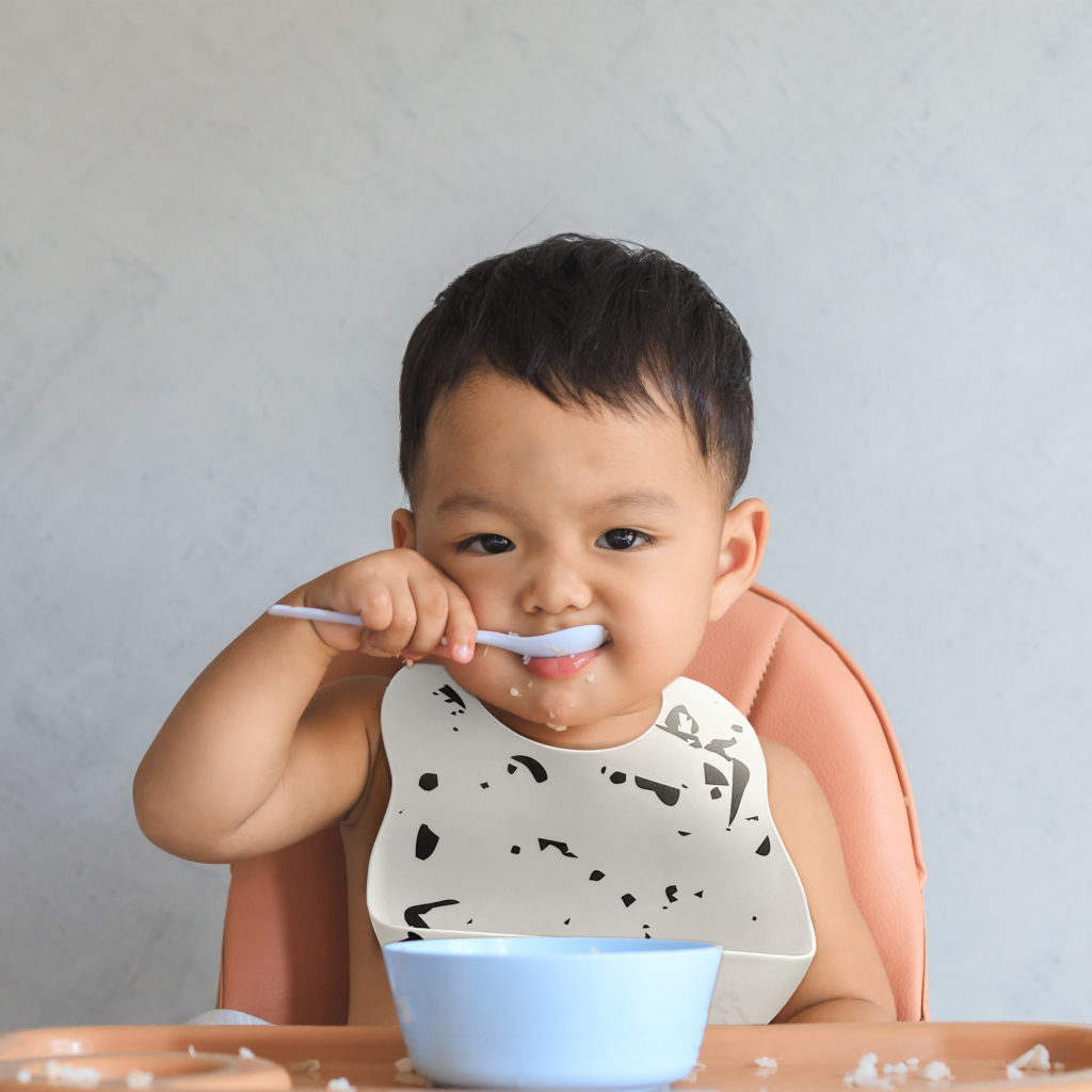 Happy infant Asian baby boy eating food by himself on baby high chair and making mess with copy space.